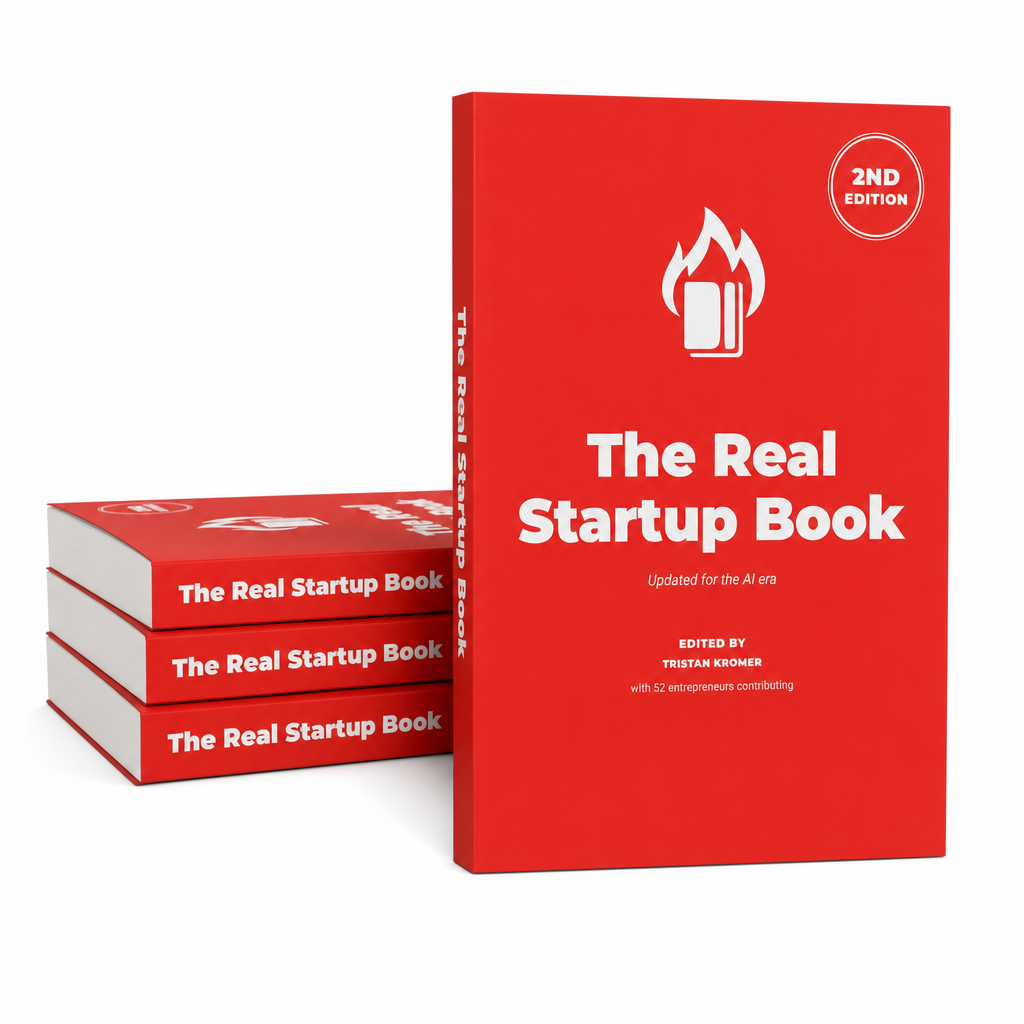 A product shot of the 2nd edition of The Real Startup Book: a red paperback edited by Tristan Kromer standing upright on a white background, with three more copies stacked horizontally behind it